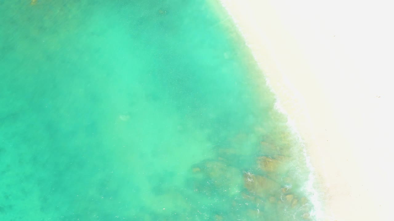 At El Tule Beach in Los Cabos, turquoise shallow water fills most of the frame beside a bright white sandy edge, with darker underwater patches and no visible people.