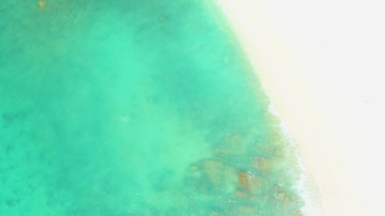 The shoreline at El Tule Beach cuts diagonally through the frame, separating bright sand from green-blue shallow water with muted underwater patches and no visible people.