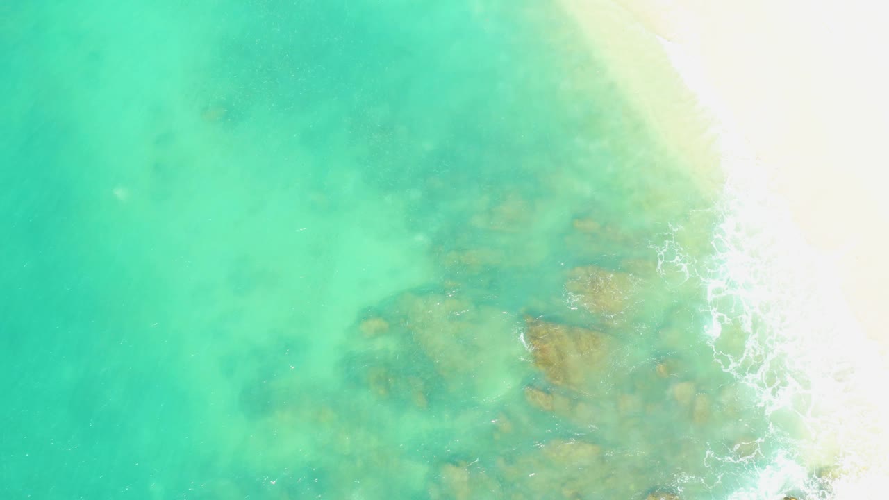 Aerial frame of clear turquoise shallows at El Tule Beach Los Cabos, with the sandy beach bright along the right edge and darker seabed forms visible under the water, no visible people.