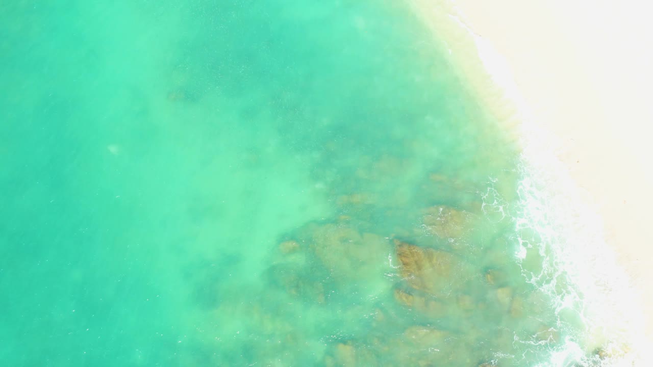 Overhead view at El Tule Beach Los Cabos showing bright sand at the right, soft foam at the waterline, and muted brown seabed patches beneath the turquoise water, with no visible people.