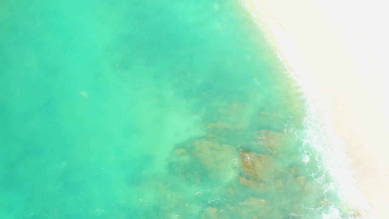 Aerial shoreline frame from El Tule Beach Los Cabos with turquoise water covering most of the view, a pale sandy strip on the right, faint foam, and no visible people.