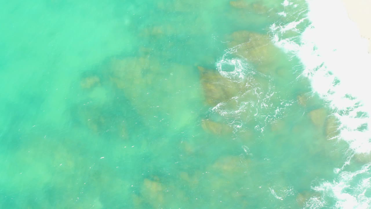 Overhead view at El Tule Beach in Los Cabos showing turquoise water over submerged brown rocks, white foam along the right side, and no visible people.
