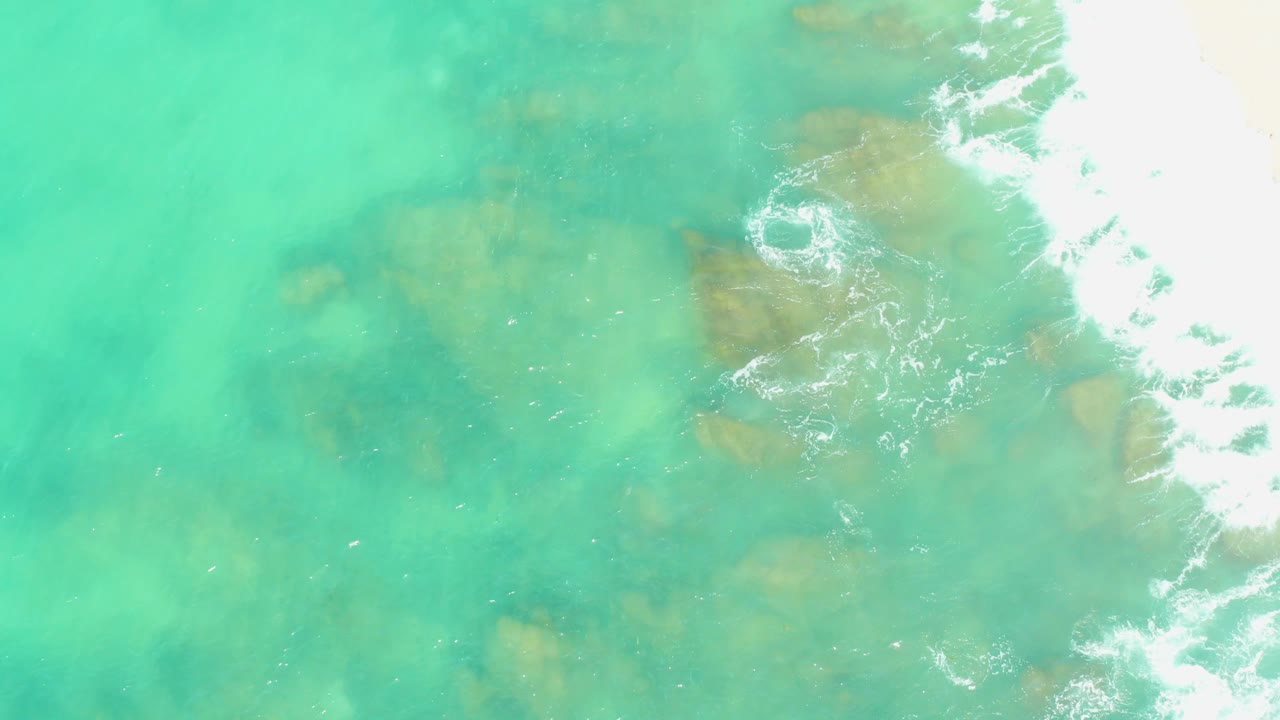 Clear green-blue water at El Tule Beach reveals scattered submerged rocks while a band of foamy surf pushes in from the right, with no visible people.