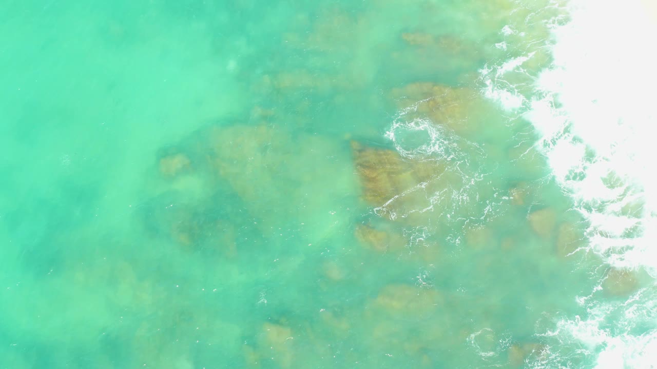 Aerial-looking frame from El Tule Beach with brown rocks under shallow turquoise water and bright white foam breaking near the right shoreline, with no visible people.