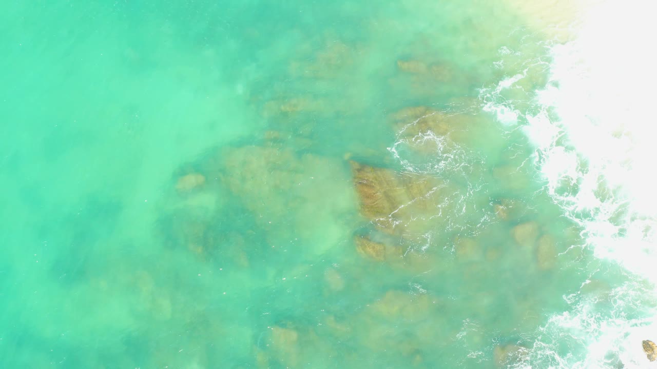Submerged rocks under turquoise water with surf foam and pale sand at the right.