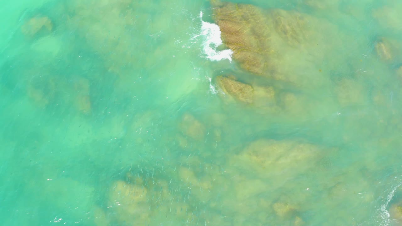 Aerial frame from El Tule Beach, Los Cabos, with submerged brown rocks under turquoise water and a brighter white foam break near the upper center, with no visible people.