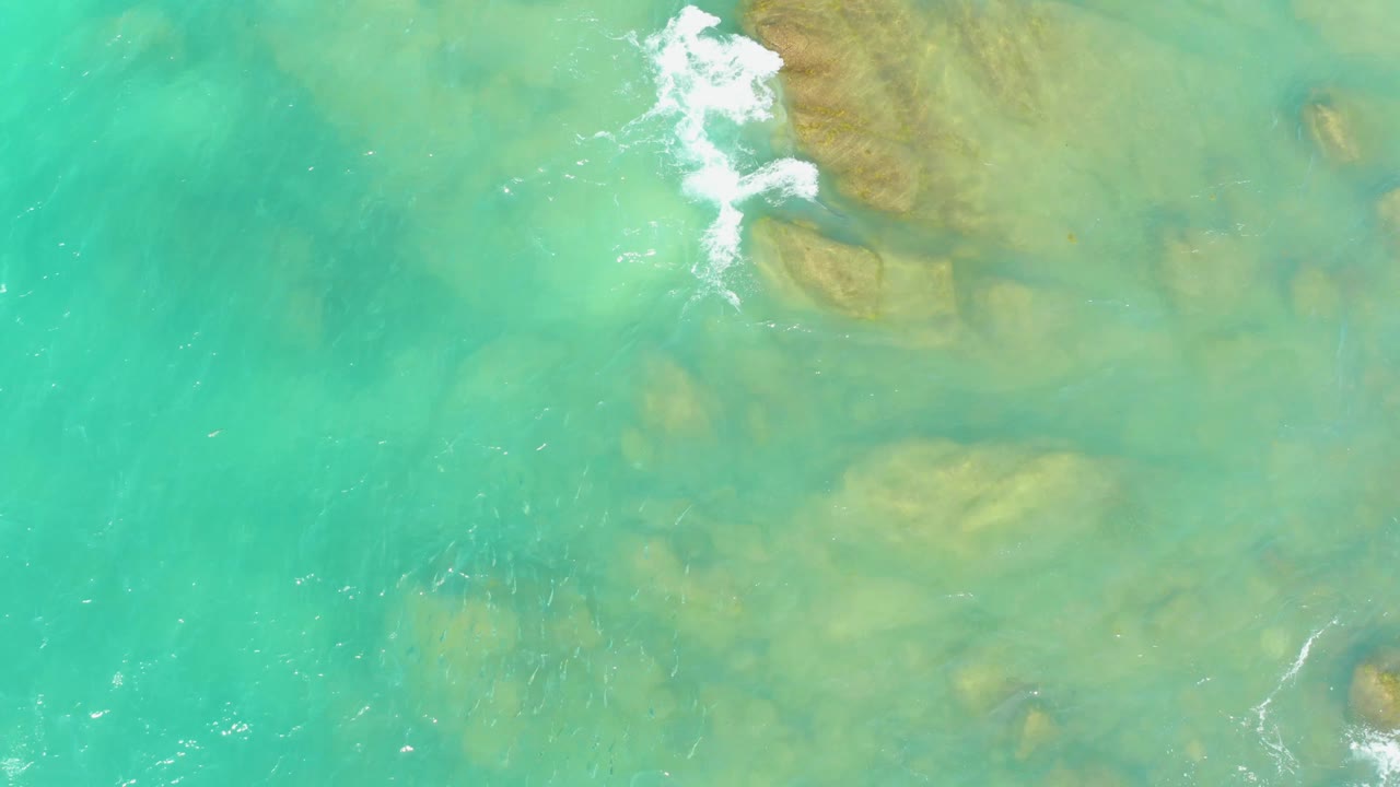 Aerial view at El Tule Beach, Los Cabos, where turquoise water covers scattered submerged rocks and a white foam patch breaks along the upper right, with no visible people.