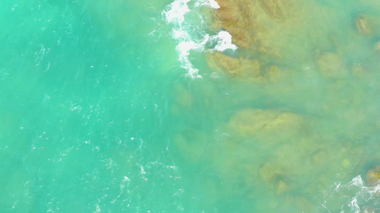 Aerial frame from El Tule Beach, Los Cabos, showing a larger white foam break spreading over submerged rocks near the top of the turquoise water, with no visible people.