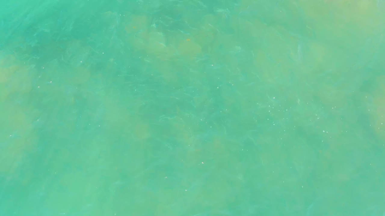 Overhead view of turquoise water at El Tule Beach Los Cabos with faint tan seabed rocks under the surface and no visible people.