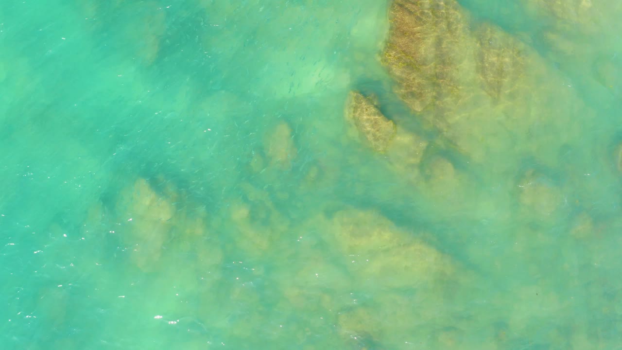Overhead view of turquoise water at El Tule Beach in Los Cabos with tan submerged rocks near the upper right and no visible people.