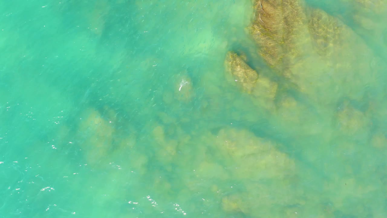Shallow turquoise water at El Tule Beach in Los Cabos reveals clustered submerged rocks near the top and diffuse seabed color below, with no visible people.
