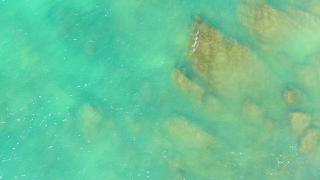 Overhead view of turquoise water at El Tule Beach Los Cabos with tan submerged rocks near the upper right and no visible people.