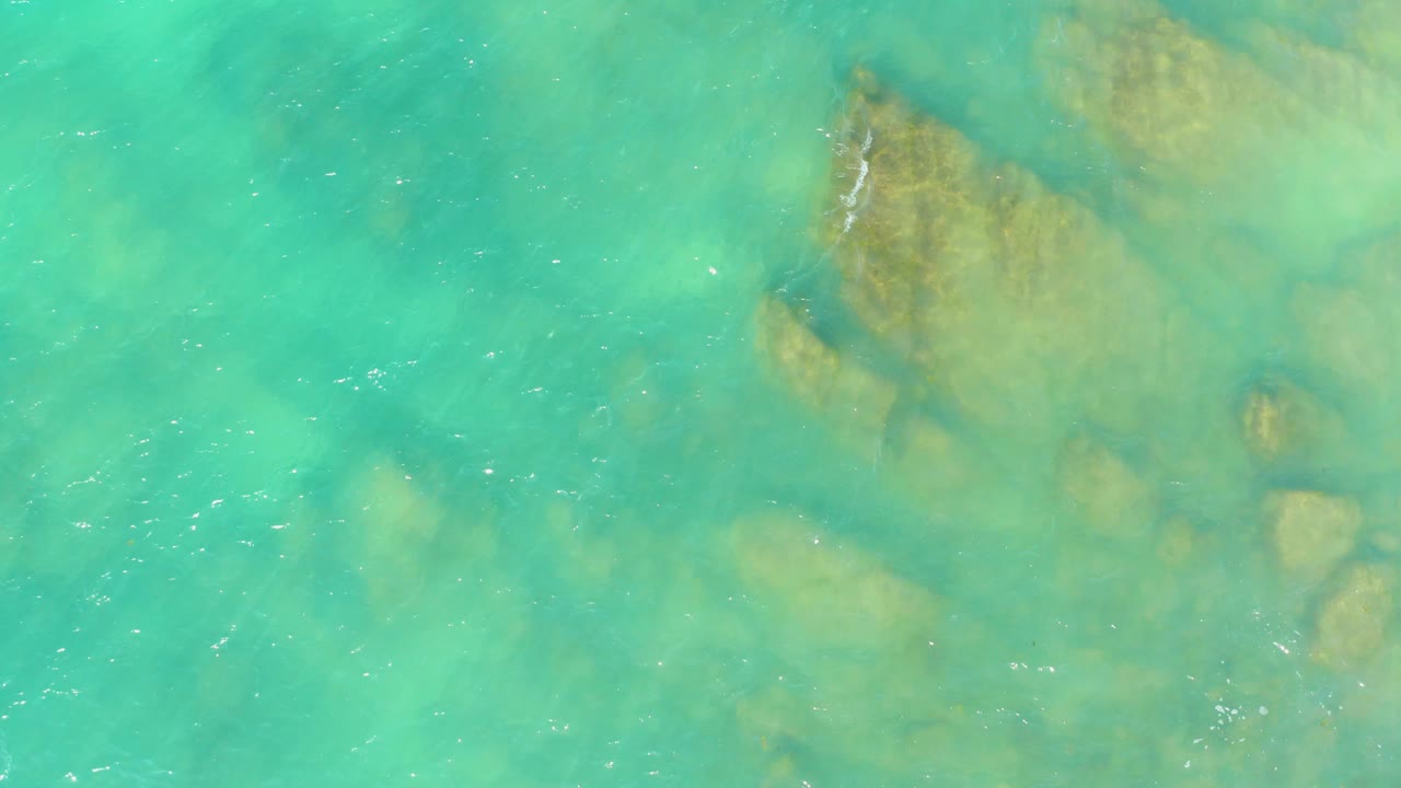 Turquoise ocean water shows submerged tan rocks through light surface ripples at El Tule Beach Los Cabos, with no visible people.