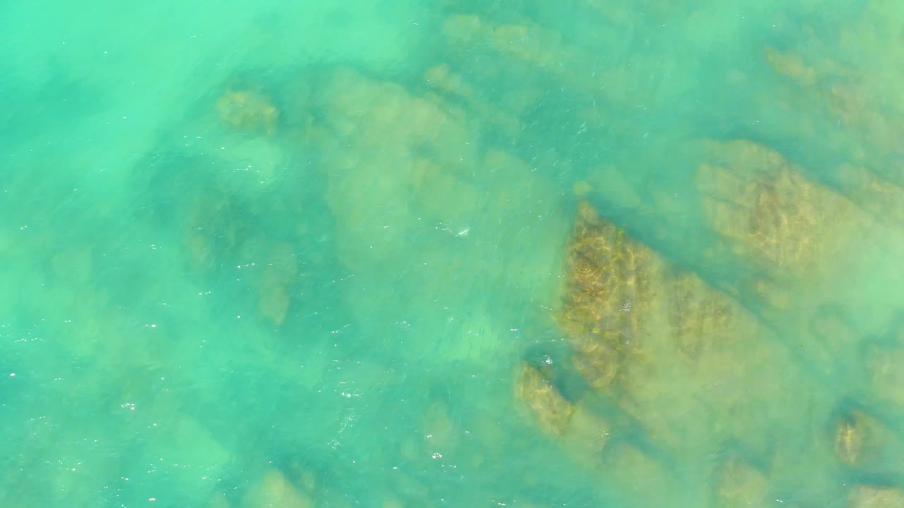 At El Tule Beach in Los Cabos, the frame shows a tan rock cluster under turquoise water, with darker underwater patches around it and no visible people.
