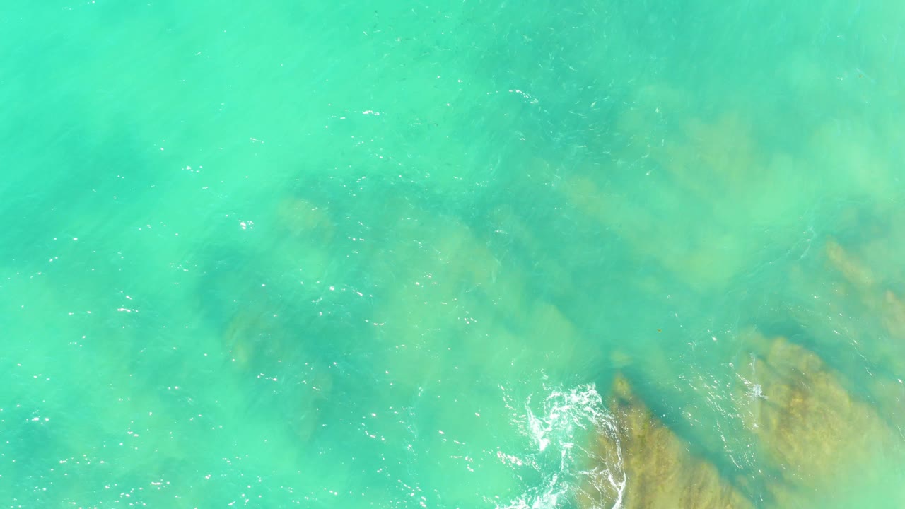 Aerial view at El Tule Beach Los Cabos showing clear turquoise water over submerged rocks, a small white foam patch near the lower center, and no visible people.