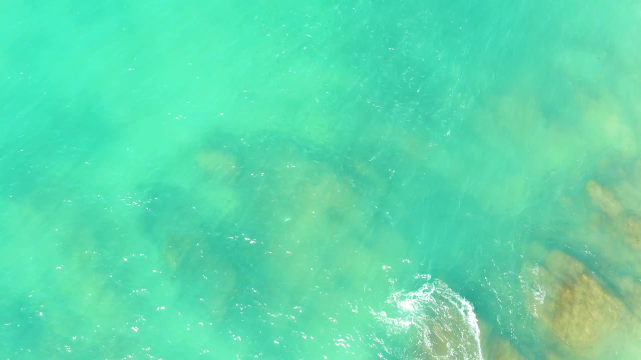 Aerial view at El Tule Beach Los Cabos showing a brighter white foam curl crossing clear turquoise water above tan underwater rocks, with no visible people.