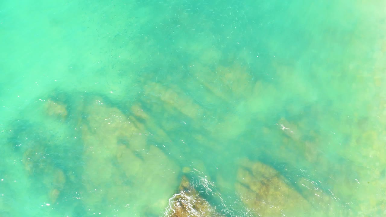 Aerial view of clear turquoise water at El Tule Beach in Los Cabos, with tan submerged rocks visible under ripples and no visible people.