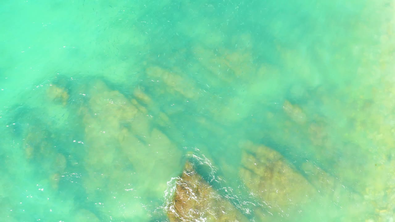 Green-blue shallow water at El Tule Beach in Los Cabos reveals submerged rock outcrops, with a small white foam break near the lower center and no visible people.
