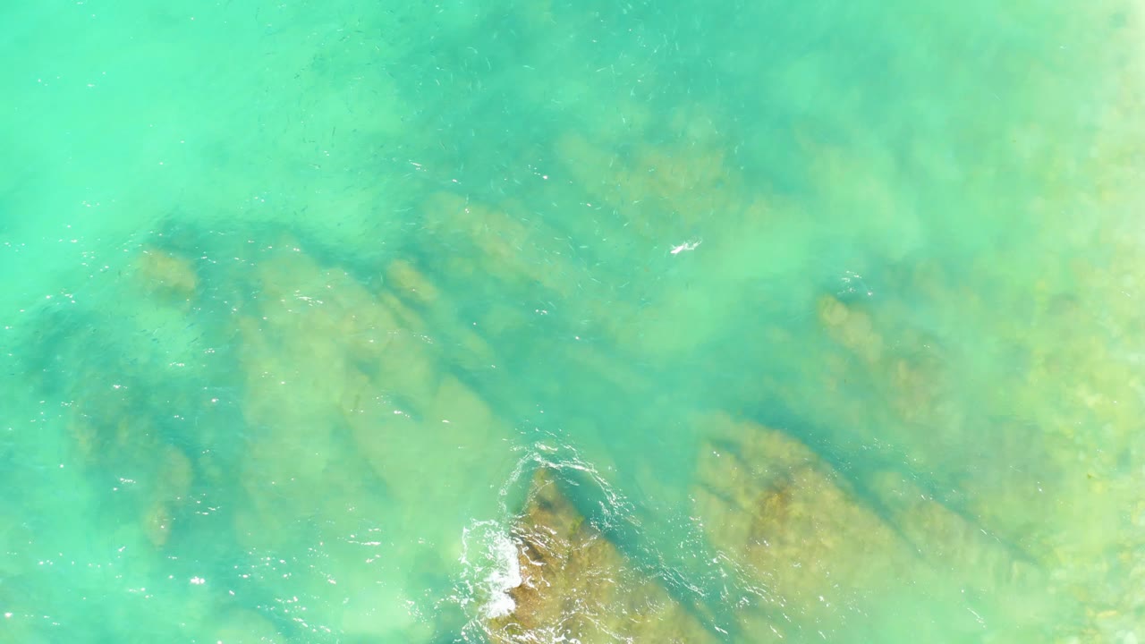 Top-down El Tule Beach water view with the rocky seabed visible through turquoise water, a foam patch touching a lower rock, and no visible people.