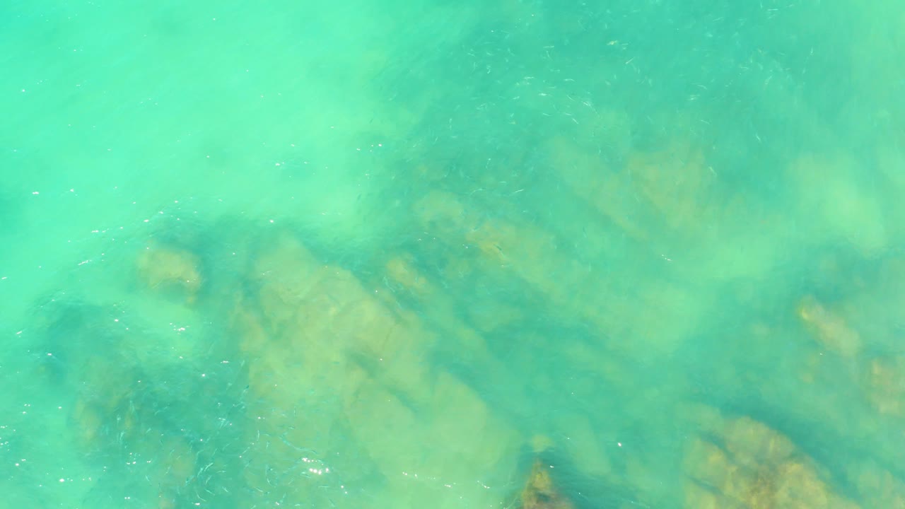 Overhead view of turquoise water at El Tule Beach Los Cabos with pale submerged rocks visible beneath soft surface ripples and no visible people.
