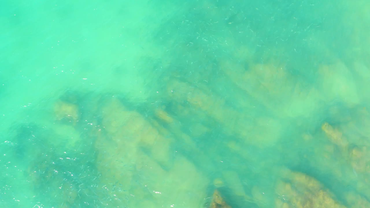 Turquoise water with faint white sparkle and tan rocky patches sits beneath an overhead view at El Tule Beach Los Cabos, with no visible people.