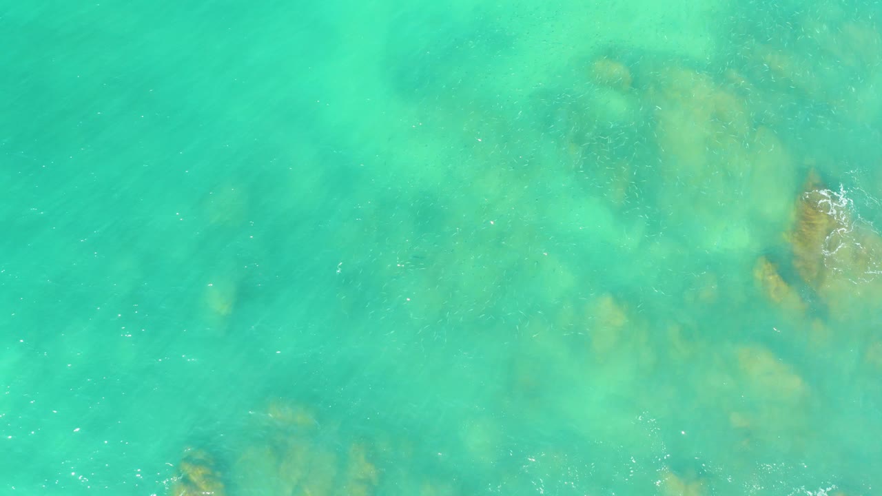 Overhead view of turquoise water at El Tule Beach Los Cabos with submerged tan rock patches near the upper and lower edges and no visible people.