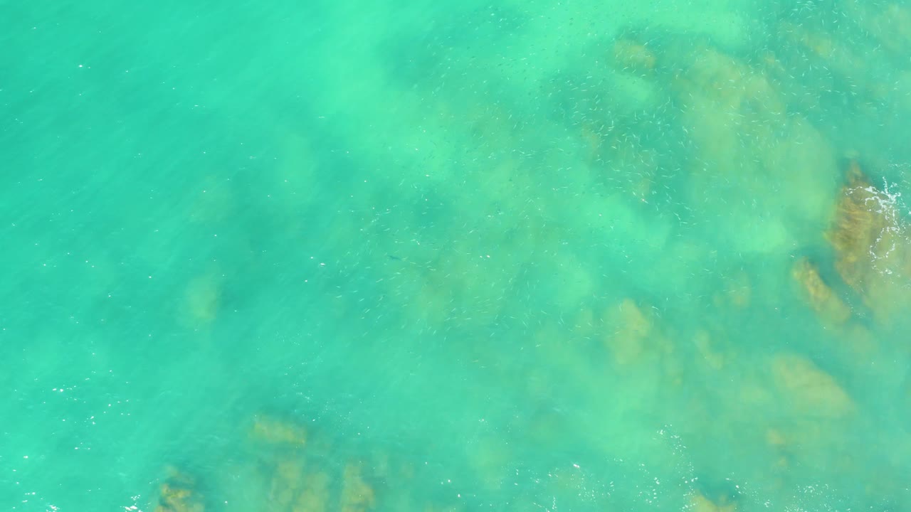 Turquoise water fills the frame at El Tule Beach Los Cabos, with darker submerged rock clusters along the right side and scattered white foam, no visible people.