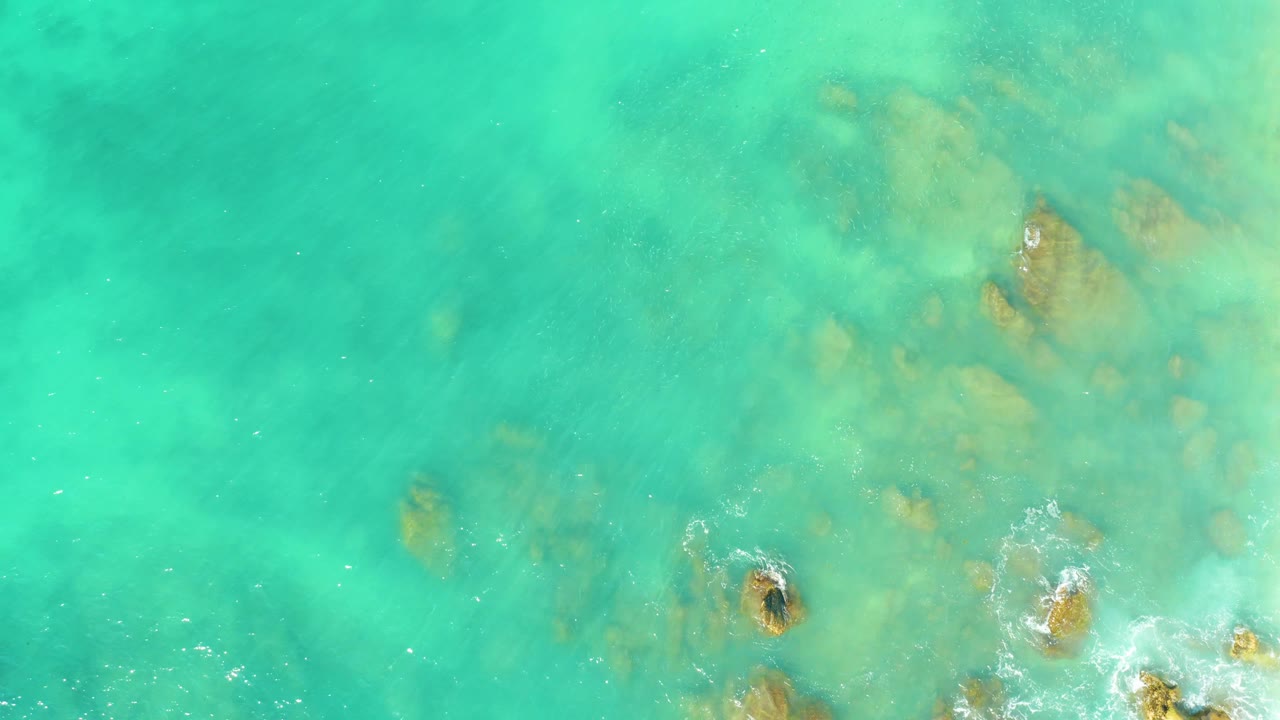 Aerial overhead frame from El Tule Beach showing green-turquoise shallow water, faint submerged rocks, and a small foam patch near the lower edge, no visible people.