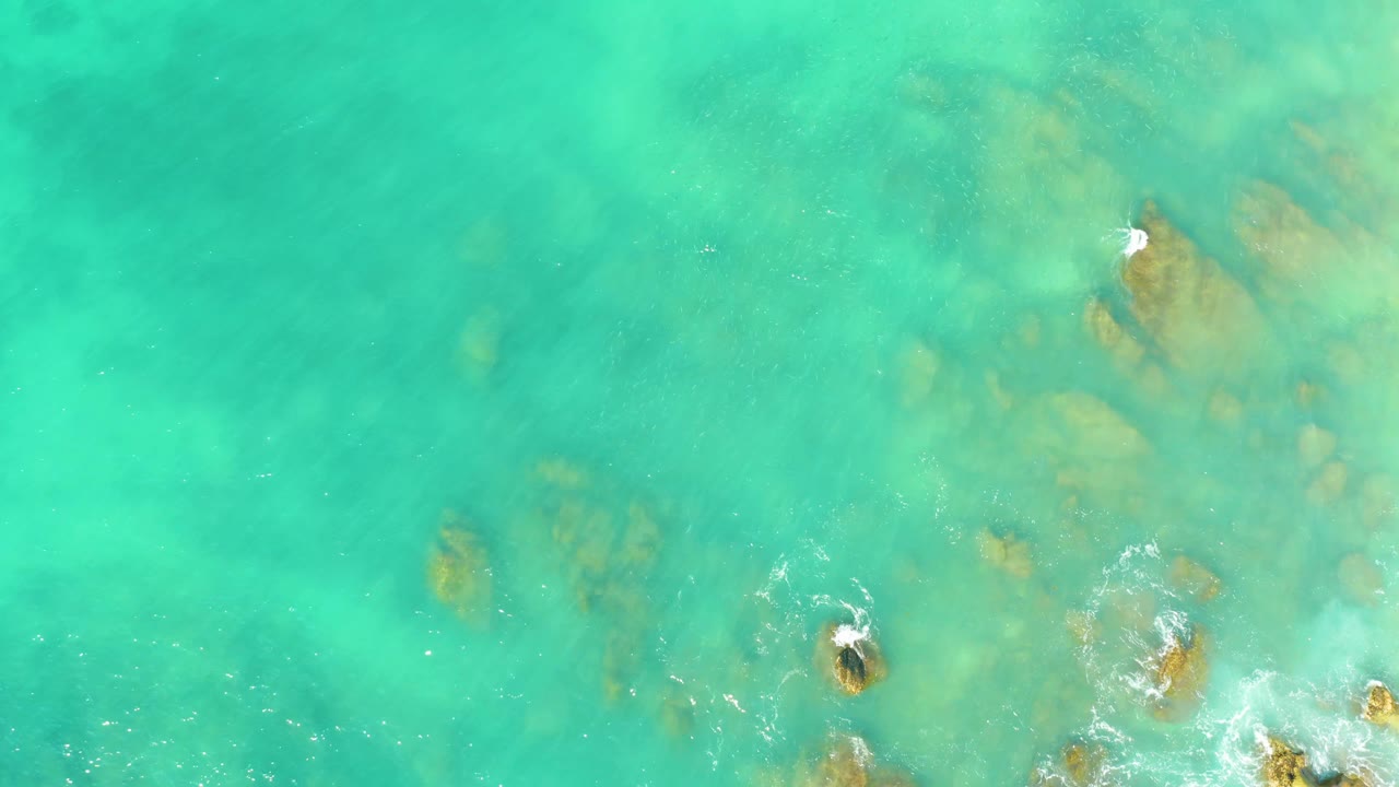 High overhead frame at El Tule Beach with turquoise water, brown submerged rocks across the right side, and white foam breaking near the frame edge, no visible people.