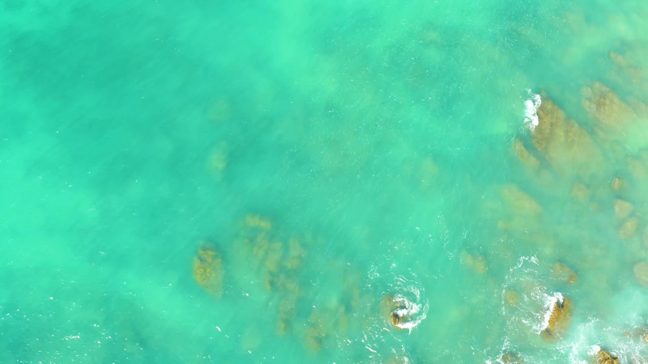 Overhead El Tule Beach frame with clear aqua water, several submerged rock shapes, and small white splash marks near the lower and right edges, no visible people.