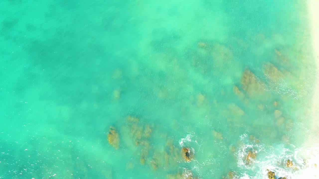 Clear green-blue water at El Tule Beach, Los Cabos, reveals clustered submerged rocks while small white foam gathers near the lower right, with no visible people.