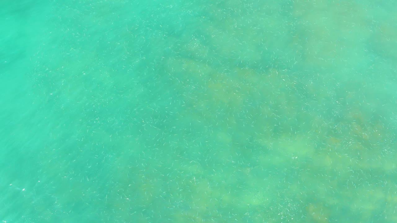 Overhead view of clear turquoise water at El Tule Beach in Los Cabos, with faint seabed shapes visible under small ripples and no visible people.