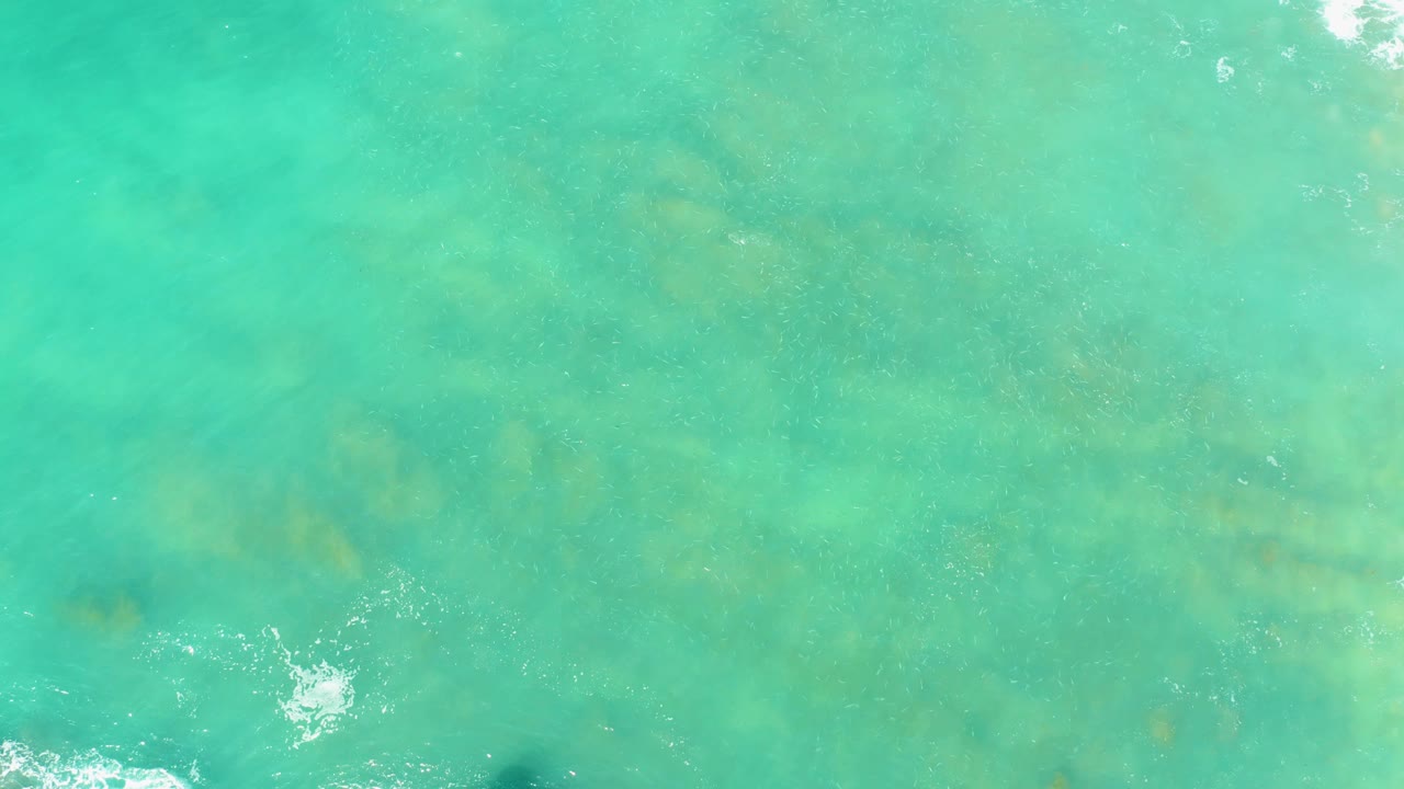 Top-down turquoise ocean water with submerged formations and a small splash near the lower left.