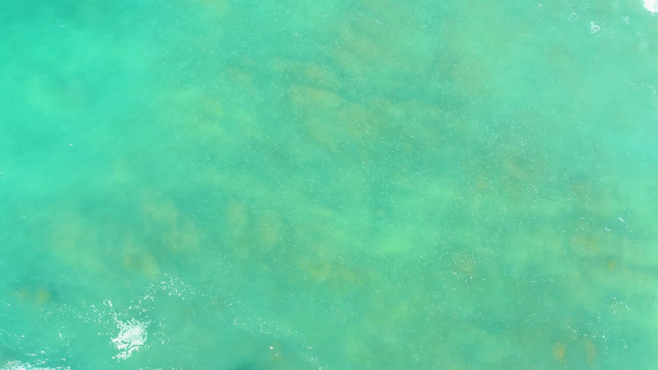 Aerial-looking view of El Tule Beach Los Cabos water, where pale turquoise surface ripples reveal underwater formations and a small white wave splash near the bottom center, no visible people.