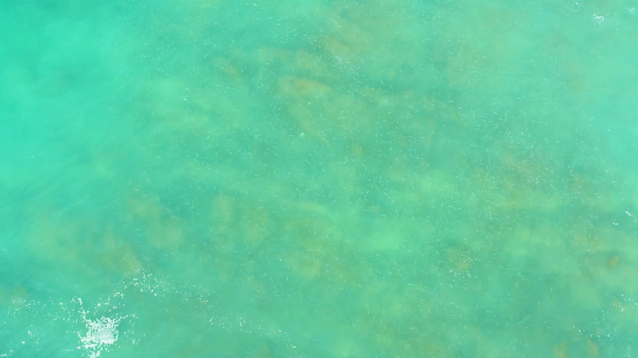 Turquoise shallow water at El Tule Beach Los Cabos shows muted green underwater formations beneath gentle ripples, with a white splash near the lower left, no visible people.