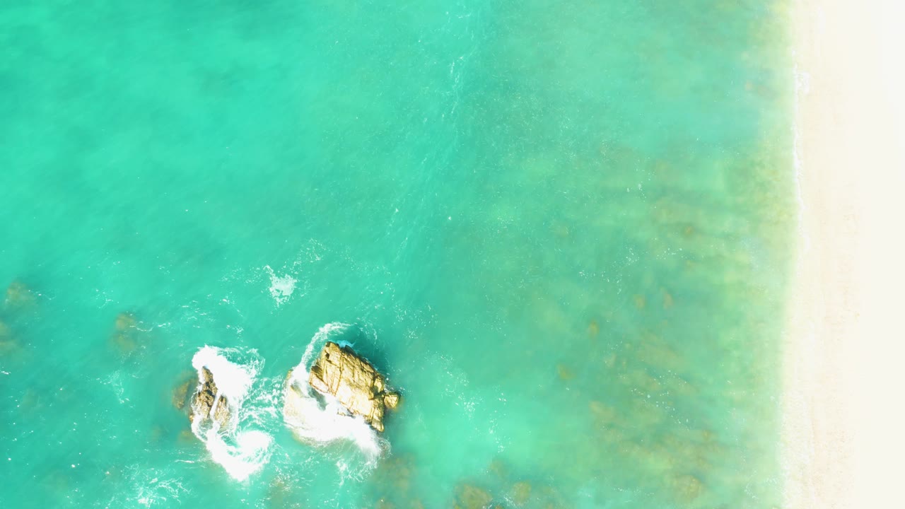 Aerial frame from El Tule Beach Los Cabos with white foam spreading around two rocks in turquoise water and a vertical strip of pale beach at right, with no visible people.