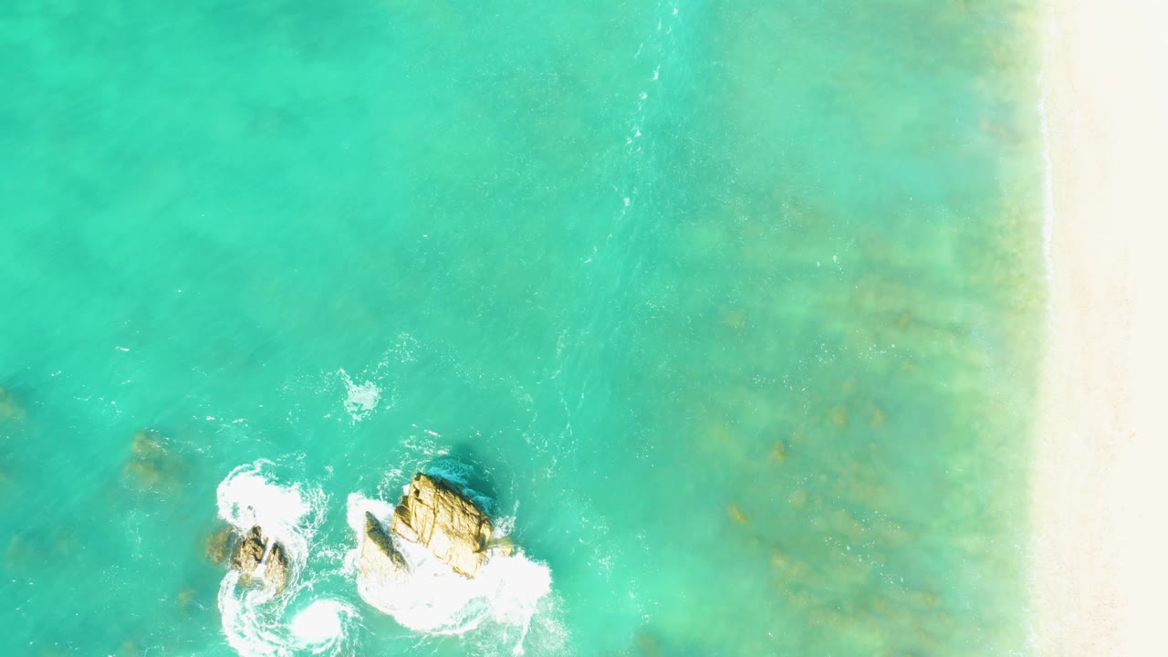 Overhead El Tule Beach Los Cabos view with the rocky outcrops lower in frame, fresh white foam around them, faint underwater patches, sand on the right, and no visible people.
