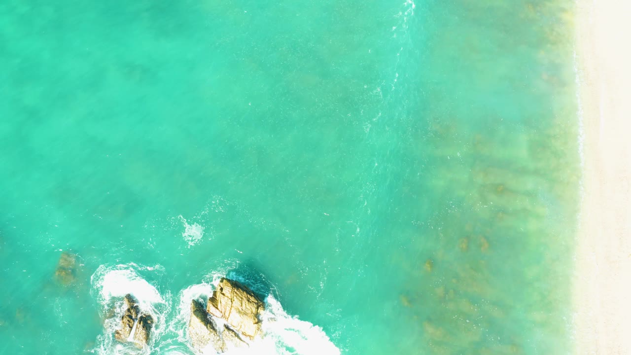 Aerial view over El Tule Beach Los Cabos with turquoise water dominating the frame, rocks partly near the bottom edge, white foam trails, right-side sand, and no visible people.
