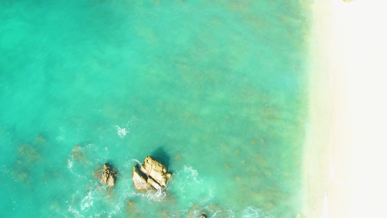 Aerial frame over El Tule Beach Los Cabos with rocks slightly below center, clear turquoise water, faint underwater patches, foam curling near the stones, and no visible people.