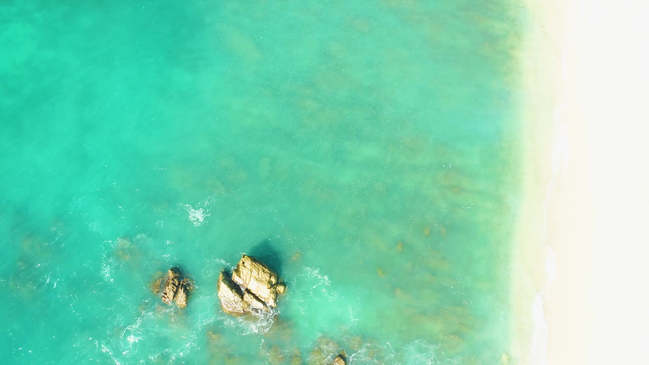 Aerial view at El Tule Beach Los Cabos with the rocks lower in frame, scattered foam to the left, clear shallow water across the view, the pale beach edge on the right, and no visible people.