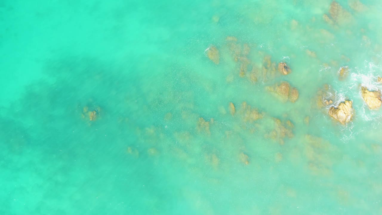 Turquoise water at El Tule Beach Los Cabos shows submerged rocks below the surface and brighter wave foam touching exposed rocks near the right edge, with no visible people.