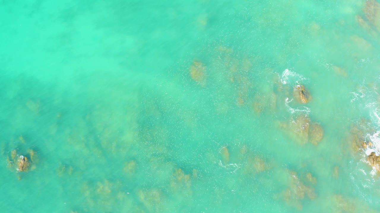Aerial frame over El Tule Beach water with pale aqua shallows, tan submerged rocks, faint seabed texture, white foam near rocks at the right edge, and no visible people.