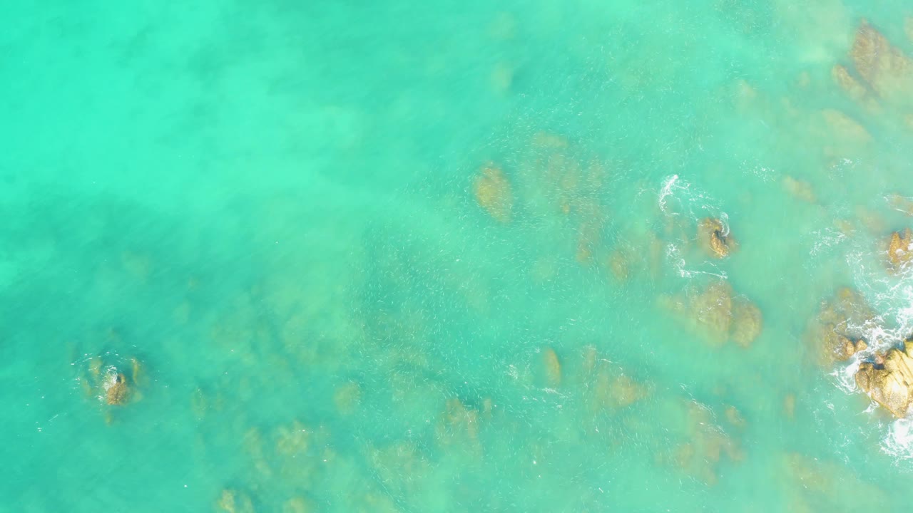 Aerial view over El Tule Beach with clear turquoise shallows, a darker submerged rock at lower left, scattered tan rocks through the center, foam at lower right, and no visible people.