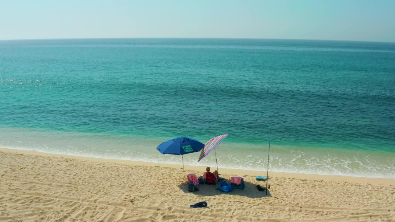 At El Tule Beach in Los Cabos, a high beach view shows seated people beneath a blue umbrella and beside a striped umbrella, with tan sand and turquoise surf behind them.