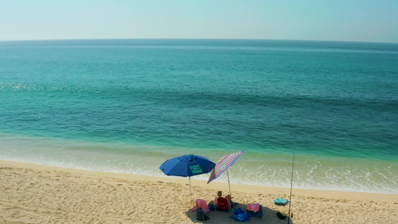 At El Tule Beach in Los Cabos, the umbrellas and seated people are pushed toward the lower right edge as gentle surf and layered blue-green water dominate the frame.