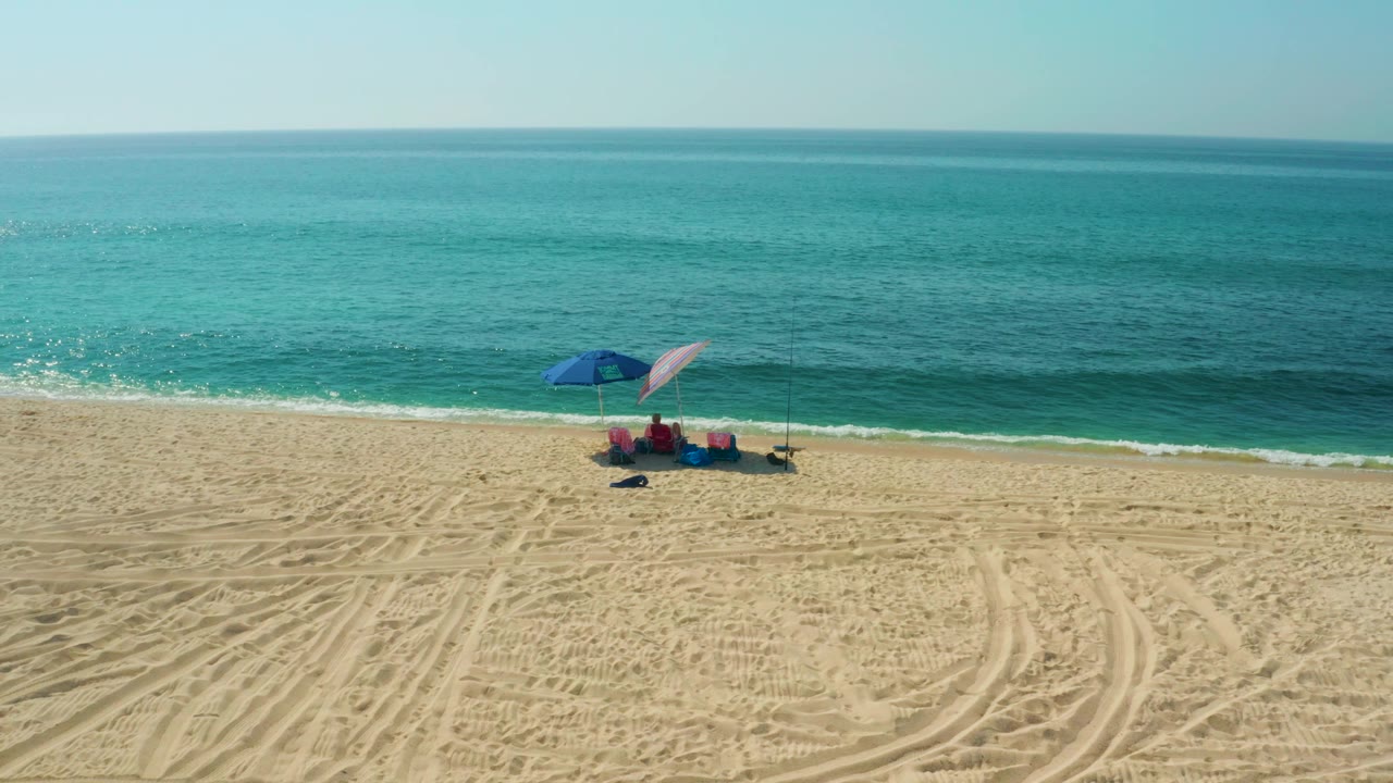 At 0:02, two umbrellas and beach gear sit low in the frame while the El Tule Beach surf forms a bright narrow band behind the setup.