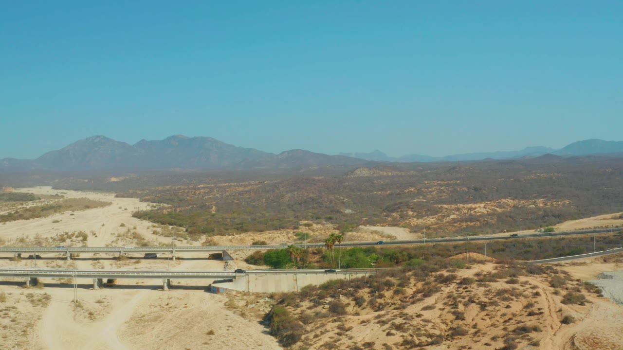 At 0:01, the El Tule Beach Los Cabos aerial view keeps the overpass low in frame, with sandy washes, scattered scrub, distant mountains, blue sky, and no visible people.