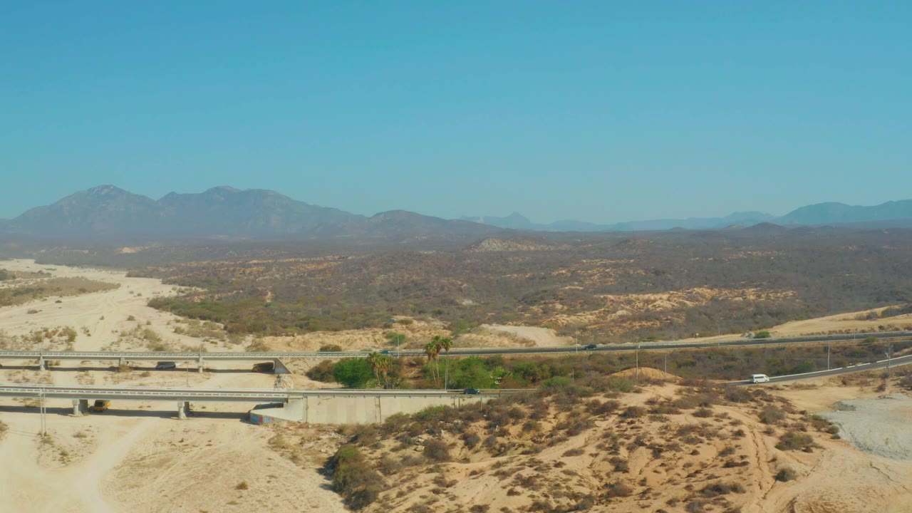 At 0:03, the El Tule Beach Los Cabos frame shows vehicles as small points on the highway, the overpass spanning sandy ground, dry brush across the valley, and no visible people.