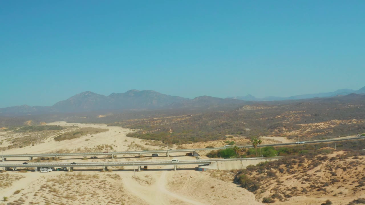 Frame three shows the highway and overpass running across the lower portion of El Tule Beach Los Cabos, with tan sand, brown scrubland, hazy mountains, clear sky, and no visible people.