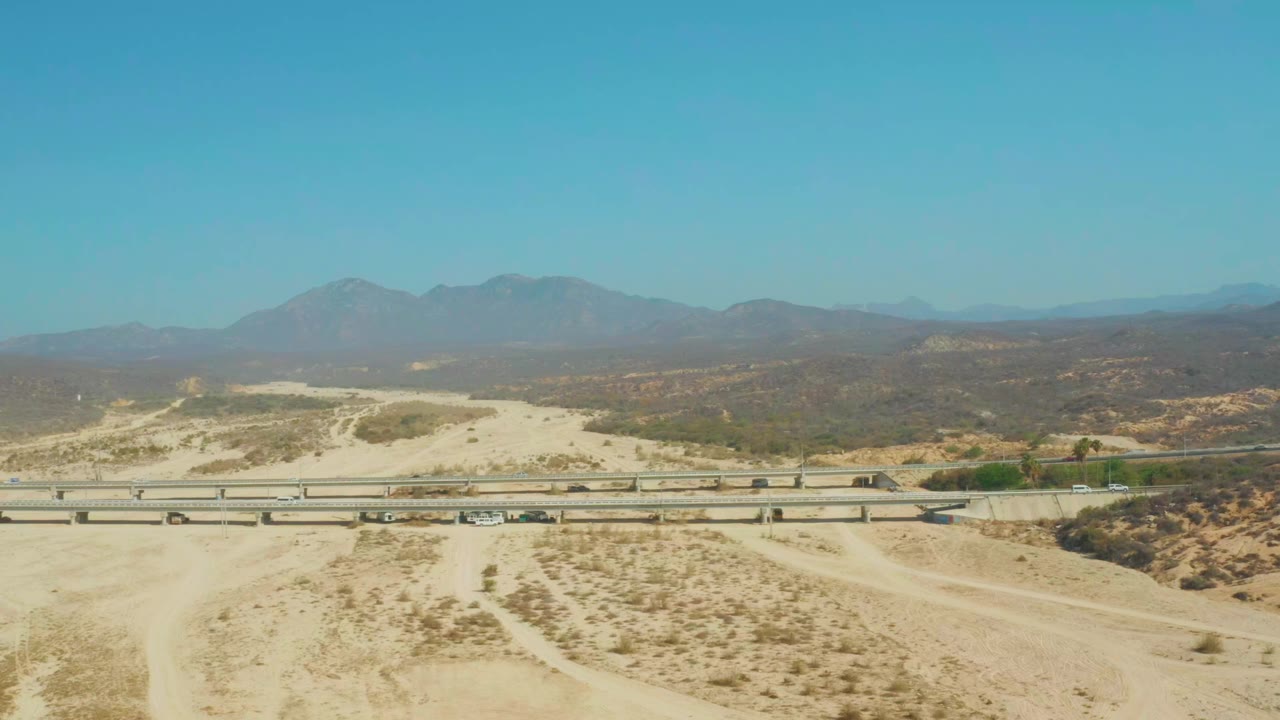 Opening aerial frame over El Tule Beach Los Cabos showing a multi-lane desert highway, pale sand, scrub patches, distant mountains, clear blue sky, and no visible people.